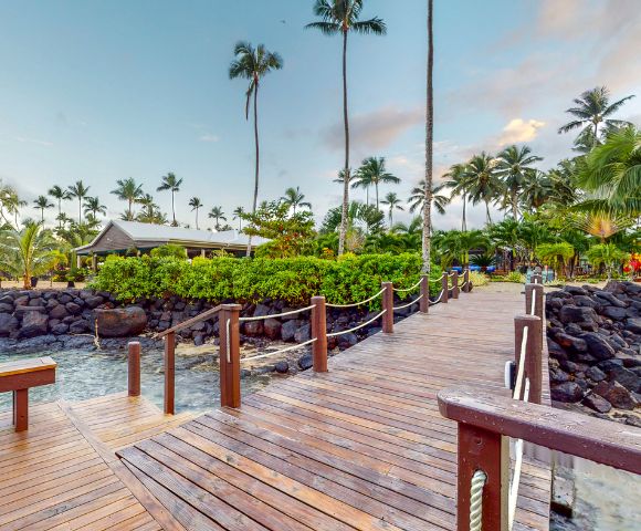 A wooden walkway leads through lush greenery and palm trees towards a serene waterside setting. A tranquil atmosphere invites relaxation.