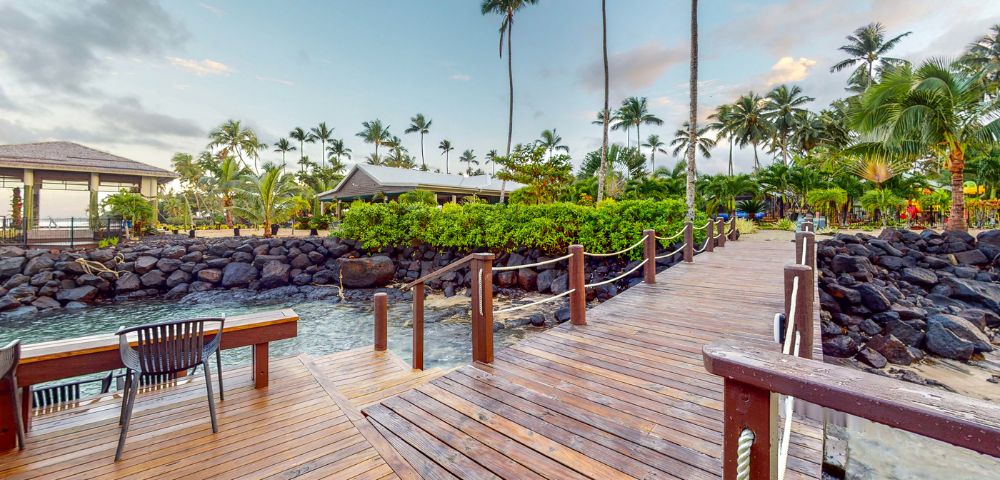 A wooden walkway leads through lush greenery and palm trees towards a serene waterside setting. A tranquil atmosphere invites relaxation.