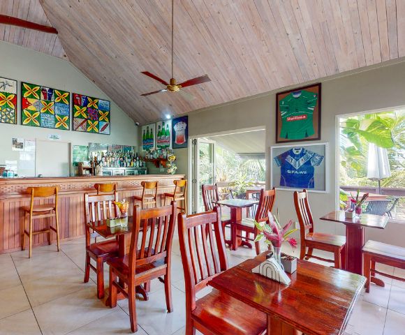 Cozy restaurant interior with wooden tables, a bar, vibrant artwork, and jerseys on display under a high, wooden ceiling.