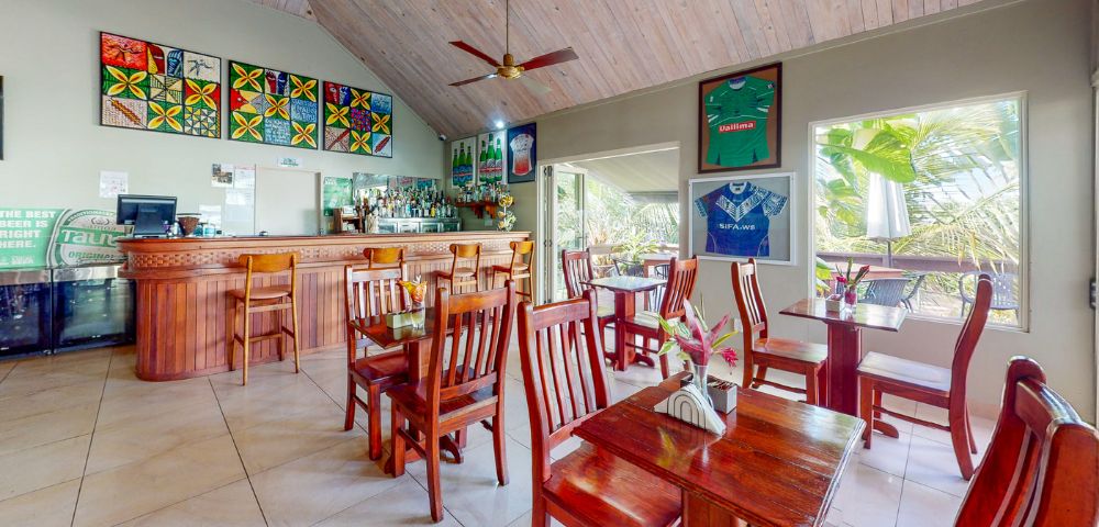 Cozy restaurant interior with wooden tables, a bar, vibrant artwork, and jerseys on display under a high, wooden ceiling.