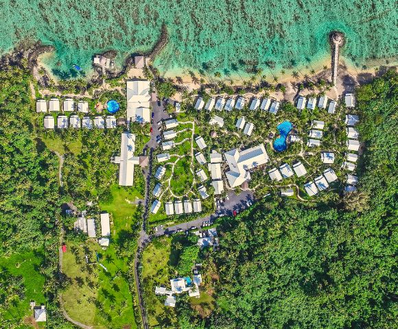 Aerial view of a tropical resort with modern bungalows, pools, and lush green landscaping by the turquoise ocean.