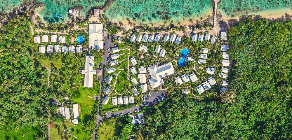 Aerial view of a tropical resort with modern bungalows, pools, and lush green landscaping by the turquoise ocean.