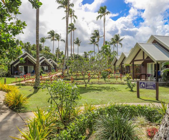Tropical resort setting with palm trees, lush gardens, and modern bungalows under a partly cloudy sky. Peaceful and inviting atmosphere.