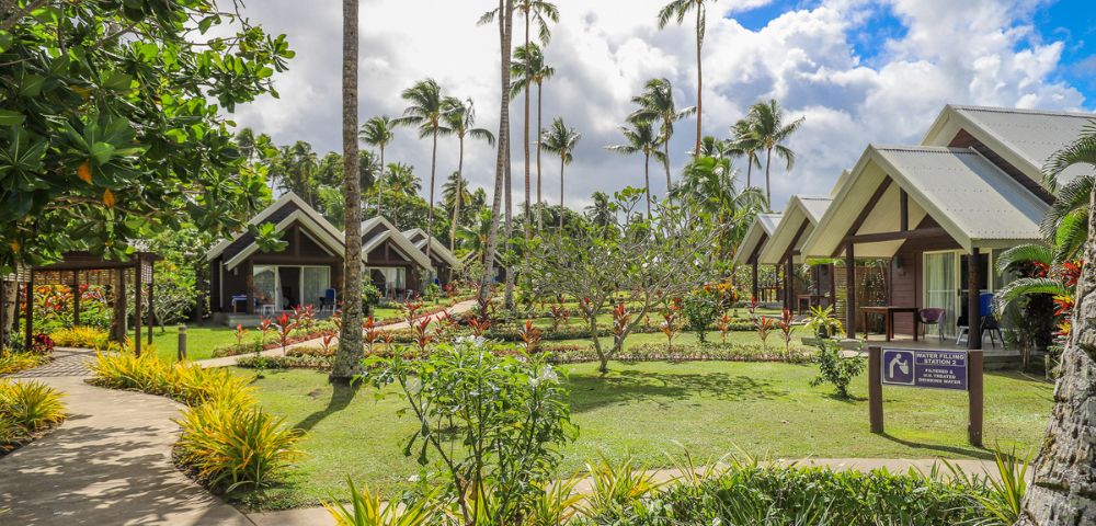 Tropical resort setting with palm trees, lush gardens, and modern bungalows under a partly cloudy sky. Peaceful and inviting atmosphere.
