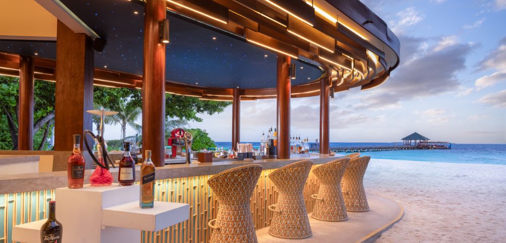 A beachside bar with woven stools, illuminated by soft lights and a starry ceiling, overlooks a serene ocean and pier at sunset.