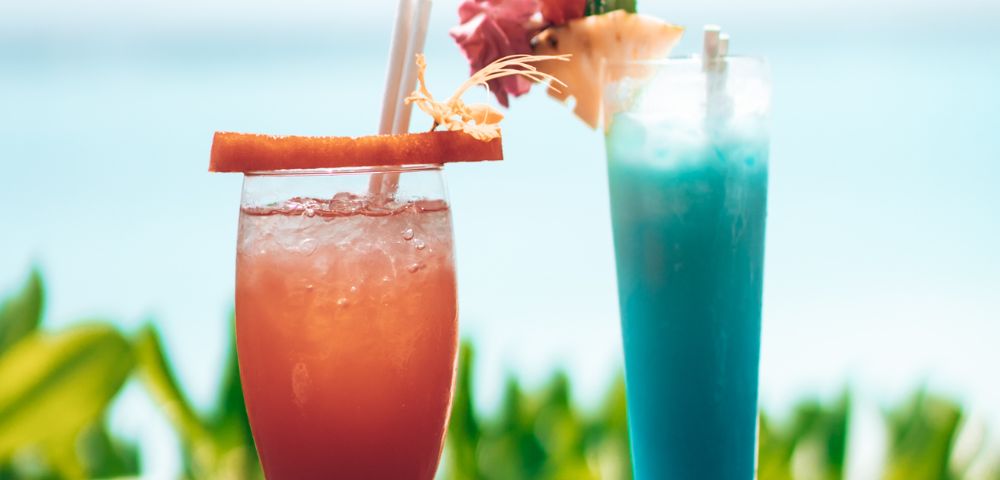 Two tropical cocktails, one red-orange and one blue, adorned with garnishes, against a backdrop of blurred greenery and ocean.