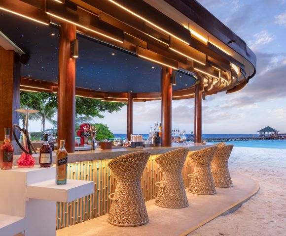 A beachside bar with woven stools, illuminated by soft lights and a starry ceiling, overlooks a serene ocean and pier at sunset.