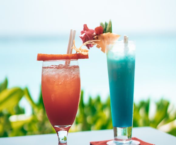 Two tropical cocktails, one red-orange and one blue, adorned with garnishes, against a backdrop of blurred greenery and ocean.