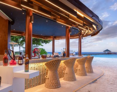 A beachside bar with woven stools, illuminated by soft lights and a starry ceiling, overlooks a serene ocean and pier at sunset.