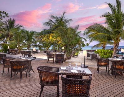 A beachside restaurant features wicker tables under palm trees, with a stunning sunset and ocean view in the background.