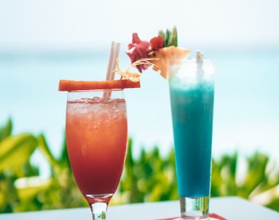 Two tropical cocktails, one red-orange and one blue, adorned with garnishes, against a backdrop of blurred greenery and ocean.