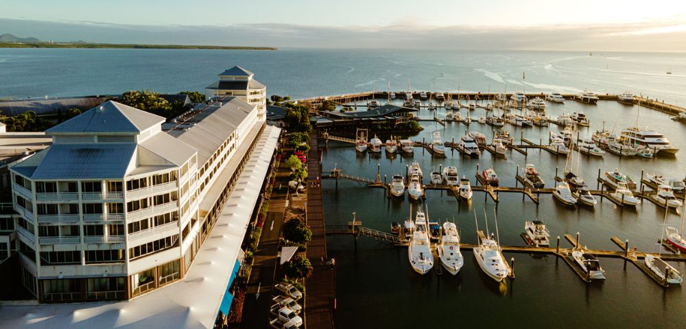 A waterfront view featuring a hotel alongside a wooden pier, with boats moored and lush greenery under a clear blue sky.