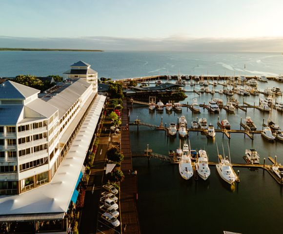 A waterfront view featuring a hotel alongside a wooden pier, with boats moored and lush greenery under a clear blue sky.