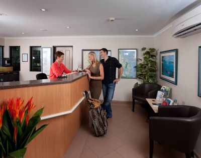 A reception area features a wooden desk, vibrant plants, and artwork on the walls, with guests engaged in conversation with staff.