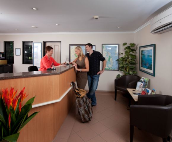 A reception area features a wooden desk, vibrant plants, and artwork on the walls, with guests engaged in conversation with staff.