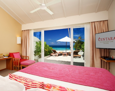 A bright hotel room with a king-size bed, featuring a balcony view of a sandy beach, turquoise water, and lounge chairs under an umbrella.