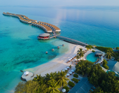 Aerial view of a luxurious beach resort in the Maldives, featuring overwater villas, clear blue waters, and lush palm trees.
