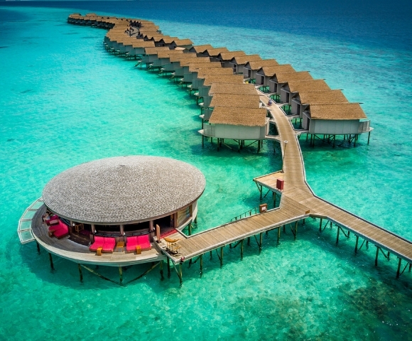 Aerial view of a luxury overwater resort with thatched-roof villas and a circular lounge, surrounded by turquoise waters.