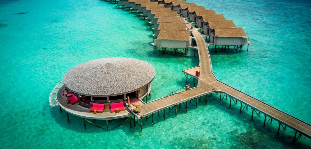 Aerial view of a luxury overwater resort with thatched-roof villas and a circular lounge, surrounded by turquoise waters.