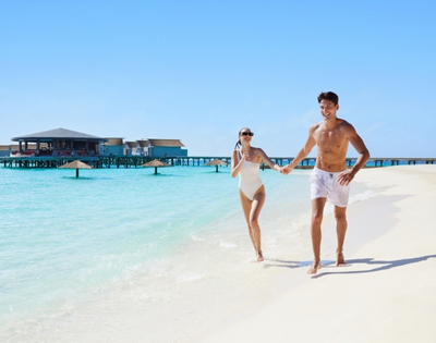 A joyful couple runs hand-in-hand along a sandy beach with clear blue water and a tropical resort in the background.