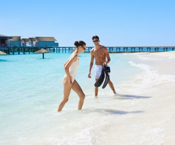 A couple walks along a sandy beach, preparing for snorkeling with fins in hand, turquoise waters and a pier in the background.