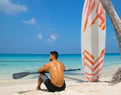 A man sits on the beach with a paddle, gazing at the ocean, beside a colorful surfboard and a palm tree. Bright blue sky above.