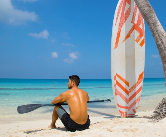 A man sits on the beach with a paddle, gazing at the ocean, beside a colorful surfboard and a palm tree. Bright blue sky above.