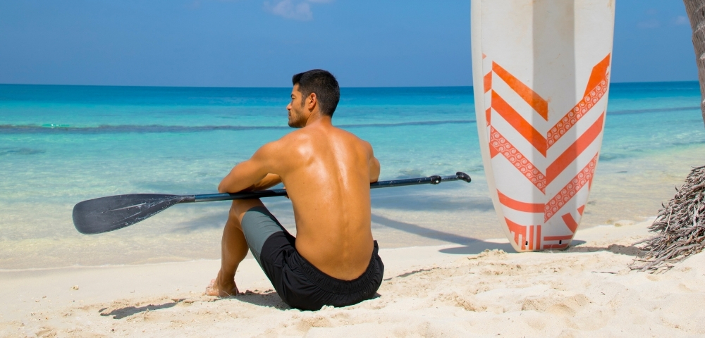 A man sits on the beach with a paddle, gazing at the ocean, beside a colorful surfboard and a palm tree. Bright blue sky above.