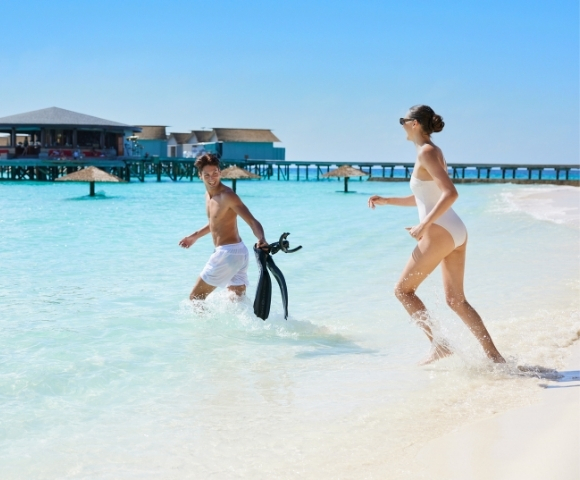 A man and woman play in shallow turquoise water at a beach, wearing swimsuits and smiling, with a pier and blue sky in the background.