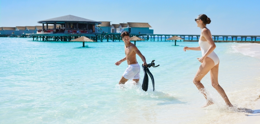 A man and woman play in shallow turquoise water at a beach, wearing swimsuits and smiling, with a pier and blue sky in the background.