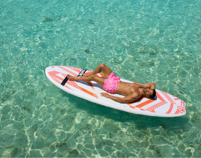 A man in pink swim trunks lounges on a paddleboard in crystal-clear turquoise water, enjoying a sunny day.