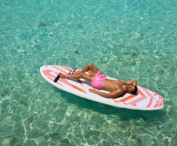A man in pink swim trunks lounges on a paddleboard in crystal-clear turquoise water, enjoying a sunny day.