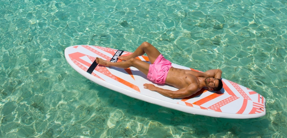 A man in pink swim trunks lounges on a paddleboard in crystal-clear turquoise water, enjoying a sunny day.