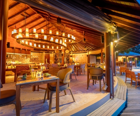 A beautifully designed beachfront restaurant with warm lighting, wooden furniture, and a circular layout, set against a twilight sky.