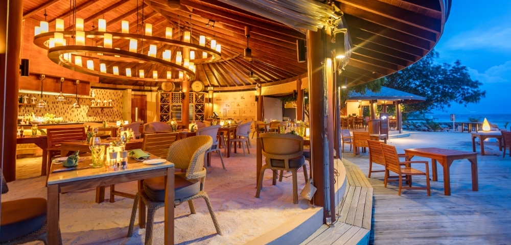 A beautifully designed beachfront restaurant with warm lighting, wooden furniture, and a circular layout, set against a twilight sky.
