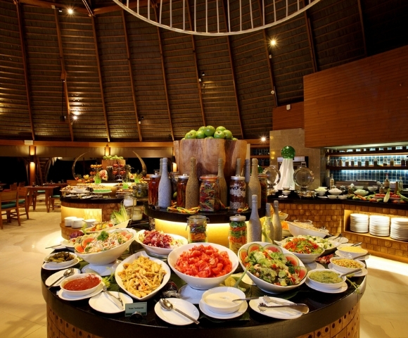 A vibrant buffet displays an array of fresh salads, sauces, and fruits in a tropical restaurant setting with wooden decor.