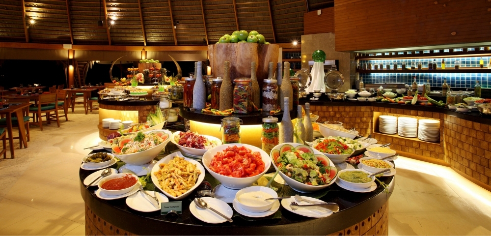 A vibrant buffet displays an array of fresh salads, sauces, and fruits in a tropical restaurant setting with wooden decor.