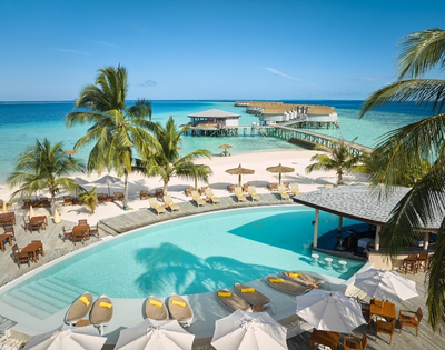 Tropical resort featuring a lagoon pool, beachside loungers, palm trees, and a pier extending into the turquoise ocean under a clear blue sky.