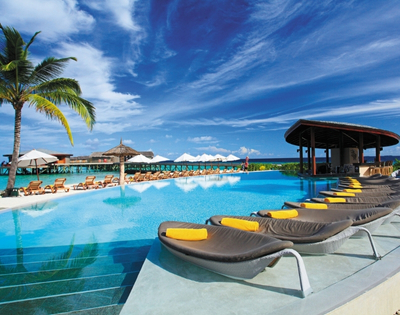 A luxurious poolside scene with loungers, palm trees, and a thatched-roof bar under a bright blue sky. Ideal for relaxation.