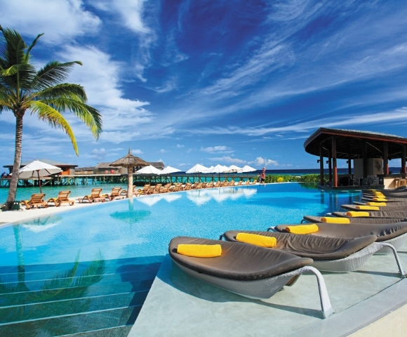 A luxurious poolside scene with loungers, palm trees, and a thatched-roof bar under a bright blue sky. Ideal for relaxation.