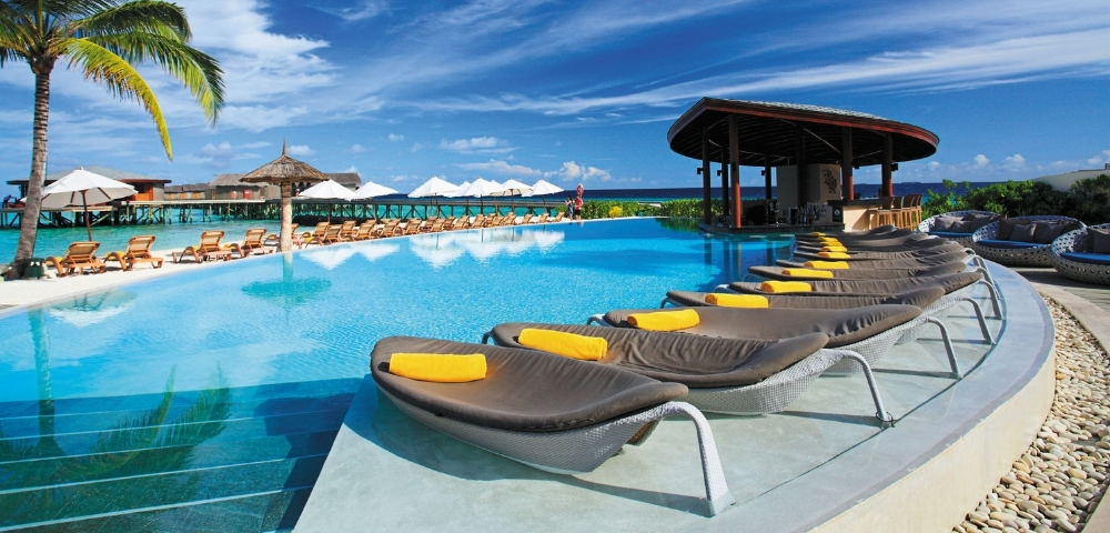 A luxurious poolside scene with loungers, palm trees, and a thatched-roof bar under a bright blue sky. Ideal for relaxation.