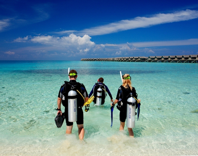 Three divers in wetsuits walking into clear turquoise water, preparing for an underwater adventure against a backdrop of blue sky and resorts.