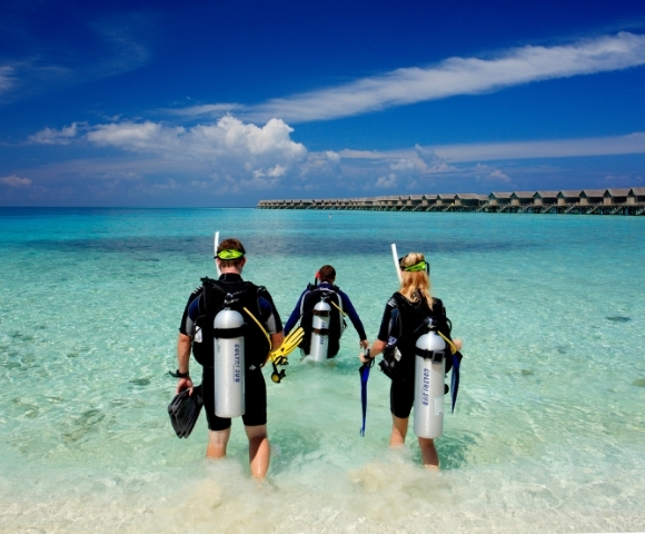 Three divers in wetsuits walking into clear turquoise water, preparing for an underwater adventure against a backdrop of blue sky and resorts.