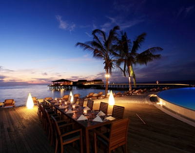 Elegant outdoor dining setup by the beach at sunset, illuminated by soft lighting, surrounded by palm trees and serene waters.