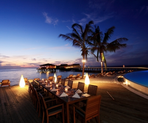 Elegant outdoor dining setup by the beach at sunset, illuminated by soft lighting, surrounded by palm trees and serene waters.