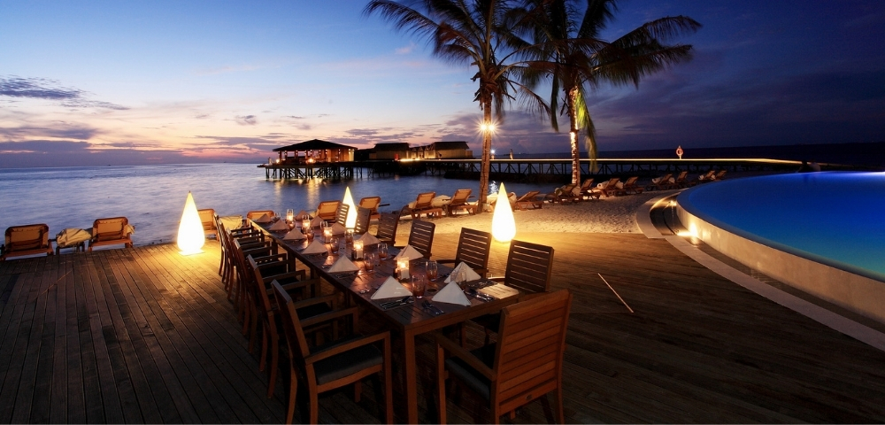 Elegant outdoor dining setup by the beach at sunset, illuminated by soft lighting, surrounded by palm trees and serene waters.