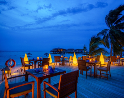 Cozy beachfront restaurant at dusk, with illuminated tables and palm trees, overlooking calm waters and a distant pier.