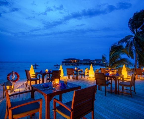 Cozy beachfront restaurant at dusk, with illuminated tables and palm trees, overlooking calm waters and a distant pier.