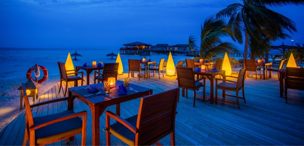 Cozy beachfront restaurant at dusk, with illuminated tables and palm trees, overlooking calm waters and a distant pier.