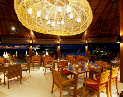 Elegant outdoor restaurant with wooden tables, vibrant blue napkins, and a large woven chandelier, illuminated against a night sky.
